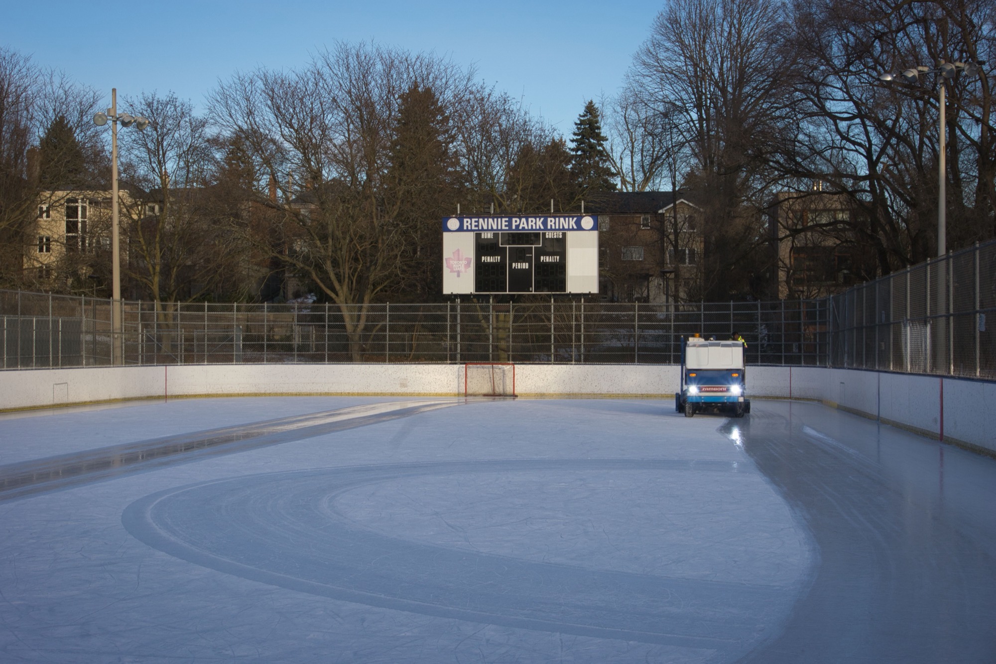 An outdoor ice rink in daylight with a resurfacing machine on the ice, no skaters present.